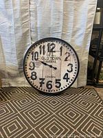 Photo showing oversized round wall clock with black hammered frame, white distressed face with large black numbers and 'OLD TOWN CLOCKS LONDON' lettering, on patterned carpet against curtain backdrop.