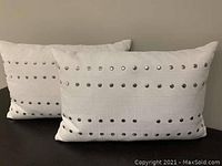 Photo showing two white rectangular throw pillows with rows of silver round metal studs, one pillow has one stud missing on the front side.