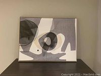Photo showing an abstract framed artwork with black, gray, and white shapes placed on a dark surface against a beige wall.
