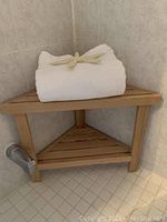 Photo of a wooden corner bench shelf in a tiled corner, with a white rolled towel on top (towel not included). The bench has two slatted shelves and three legs.
