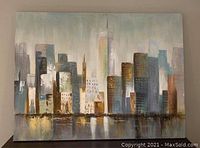 Photo shows abstract cityscape painting featuring various buildings in soft neutral and metallic colors with reflection on water below.