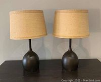 Pair of table lamps with dark rounded bases and beige fabric shades, sitting on a dark surface against a light background.
