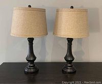 Photo of a pair of black table lamps with beige drum fabric shades, positioned side by side on a flat surface with a neutral background.