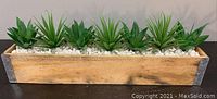 Wooden rectangular planter box with metal reinforcement corners, filled with white pebbles and seven artificial succulent plants lined up inside.