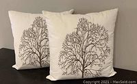 Photo of two white throw pillows with brown tree branch design, placed side by side on dark surface against a neutral background.