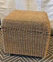 Wicker ottoman square shape with woven pattern, showing front and one side, on a patterned carpet.