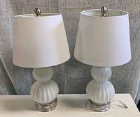 Pair of white glass table lamps with white drum shades on a white surface in front of grey fabric backdrop, showing front and side angles.