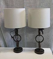 Photo showing full front view of both lamps, highlighting their matching dark metal bases with circular link design and white fabric drum shades.