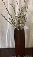 Photo shows a tall rectangular dark-stained wooden vase containing artificial pussy willow branches with white catkins against a textured light-colored background.