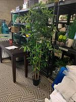 Full height view of the tall faux bamboo plant in black pot placed indoors beside a table with shelving in the background.