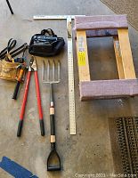Complete view of all items arranged on floor including toolbelt with hammer and pliers, pitch fork, loppers, furniture dolly, large T-square, and electric sander in case.