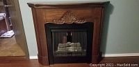 Front view of the walnut-stained decorative faux fireplace showing carved detail above firebox and black mesh screen with faux logs inside.