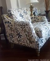 Image shows a beige linen-like upholstered accent chair with green leaf designs, nail head trim around edges, and turned front wooden legs in antique weathered grey finish. Chair cushion and back cushions have the same leaf pattern upholstery.