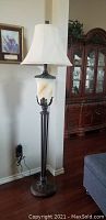 Full view of the tall floor lamp showing the cream bell shade, frosted urn, and metal base in a living room setting.