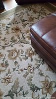 Photo showing part of the floral patterned area rug with ivory background and green, beige, gold flowers near a brown leather furniture piece.