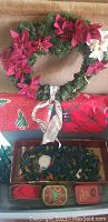 Photo showing green Christmas wreath decorated with red and white poinsettias, fabric festive table cloths, vintage light strands in a box and two red tin boxes.