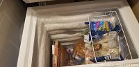 Top-down view of the open white chest freezer showing the insulated interior, some frozen food items, and a removable wire basket inside.
