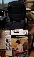 BackMasseur black paddded back massager and seat cover with back brace strap. Includes original box with product branding and features visible. Ergonomic hand control with illuminated buttons connected.