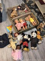 Vintage plush toys including Mickey Mouse, Raggedy Ann, baby dolls, and other assorted plush toys displayed with a carrying suitcase.