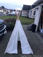White fabric tipi tent standing outdoors on pavement in front of a house. The tent is supported by wooden poles crossing at the top, with the entrance flap open.