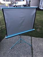 Full view of the Da-Lite Silver-Lite projector screen on its tripod stand set outdoors on a paved surface with a grassy background.