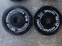 Photo of 2.5 pound iron weight plates in black with white lettering on concrete floor, clear readable markings 'WASHINGTONIAN NM TACOMA 2 1/2 LBS'