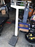 Full view of the vintage mechanical standing weight scale standing on a cement floor among other garage items