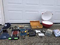 Wide shot showing majority of lot items arranged on ground outside garage - including cooler, hats, tackle box, golf balls, paddles, tool pouch, bartending item