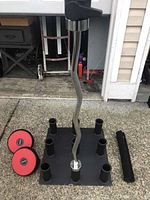 Top view of the solid 9-slot vertical Olympic bar holder and Olympic curl bar with collars, two dumbbells, and a squat pad visible to the sides.