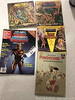 Photo showing four He-Man/Masters of the Universe magazines and one Walt Disney Pinocchio book, all with visible wear and tear.