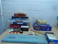 Overview of the stacked games including Monopoly, Backgammon, Pachisi, Booby-Trap, and Kalah among others, laid out on a table against a brick wall.