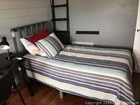 Single gray metal bed frame with slatted headboard and side rails, set up in room with hardwood floor. Bed made up with striped blue, white and red bedding and pillows.