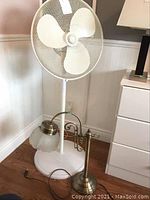 Full view of white standing fan with three blades and mesh grille, next to metal desk lamp base