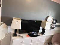 Two matching modern table lamps placed on white drawer unit, also visible a flat-screen TV behind and other room furnishings. Lamps have white square shades and dark rectangular bases with silver metal trim.