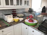 Wide shot showing kitchen countertop with toaster, kettle, juicer, mixing bowls, plastic trays and storage containers.
