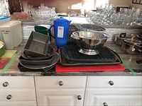 Photo showing stacked metal baking pans including round cake pans, rectangular loaf pan, and cookie sheets. Red cutting board and metal colander placed on counter with pitchers in background.