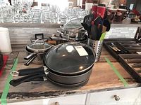 View of the Bialetti non-stick fry pan with glass lid and black handle in front, with Lagostina pots and lids stacked behind and metal utensil holder with various utensils to the right.