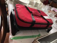 Hamilton Beach electric grill flat on table with red insulated food carrier bag on top.
