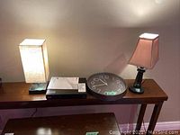 Photo of both lamps lit on a wooden console table alongside LG Blu-Ray player with manual and a round clock.