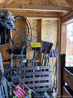 Photo showing various garden tools leaning against a wooden wall including rakes, shovel, garden broom, garden hose coiled on reel, and a hula hoop.