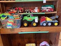 Photo of various toy vehicles including a green dump truck, red and yellow tow truck, and assorted other toy vehicles, along with a bin filled with colorful flexible plastic track pieces.