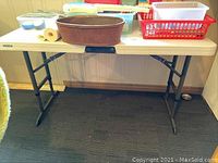 View of full folding table showing rectangular white resin top with various items on it and black folding metal legs.