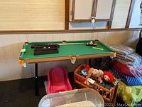 Full view of mini pool table with folding metal legs, green felt surface, pool cues, and accessories visible on top, surrounded by other household items.