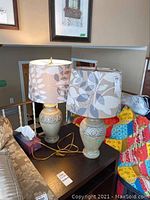 Pair of beige ceramic table lamps with carved ornamental scroll designs on the bases, topped with drum fabric shades featuring a taupe and cream leaf pattern. Lamps placed on a black table next to a couch and other household items.