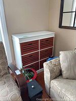 Front view of white storage unit with six brown wooden slide-out crates between two chairs, plant and red pot visible in the foreground.