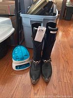 Pair of black cowboy boots with toe caps and harness straps shown next to the Conair nail care station on hardwood floor with bin in background.