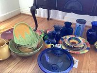 Wide shot showing mixed ceramics and blue glass items on floor including corn platter, salad bowls, sunflower bowl, pitchers, and blue glass vases.