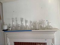 Wide view showing clear glass decanters and pitchers lined up on mantelpiece, totaling 11 decanters and 2 pitchers of varying heights and shapes