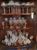 Wooden cabinet shelves with full assortment of stemware and barware: top shelf holds 7 large balloon-style wine glasses; middle displays mixed smaller glasses; bottom shows salt & pepper shaker, butter rests, small glasses and paperweights.
