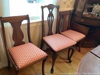 Three wood dining chairs with medium brown finish, two with curved cutout backs and upholstered orange geometric patterned seats, and one with carved wood back detailing.